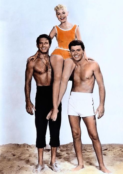 A teen girl, clad in an orange one-piece swimsuit and with her blonde hair pulled into a short ponytail, sits on the shoulders of two men who are standing on what appears to be a sandy beach. The man on the left is visibly older and wearing dark pants with the legs rolled up, while the one on the right looks only a year or two older than the girl and is wearing white swim trunks. All three are smiling widely.